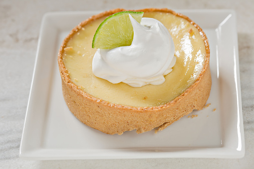 A high angle shot of a small square plate with a personal sized key lime cheesecake tart topped with fresh whipped cream and a wedge of fresh lime. Shot on a white marble surface.