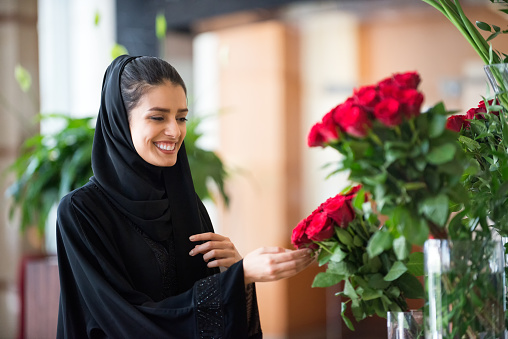 iStockalypse Dubai.  An attractive young Emirati, Middle Eastern woman florist wearing a black abaya and hijab touches a bunch of long stemmed red roses in a flower arrangement displayed in the lobby of a modern hotel.  Dubai, U.A.E., Middle East