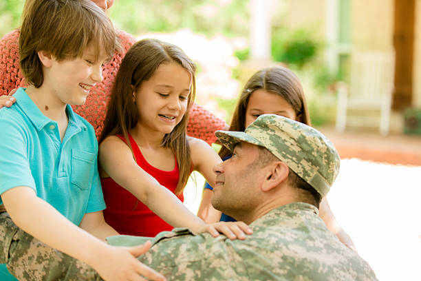 American military veteran welcomed home by family. Mom, children. Home. Handsome U.S.A. military soldier being welcomed home by his family. Wife, two little girls and one boy children. Home in background. He wears his military uniform and is excited to see his family. The children and wife eagerly give him a big hug. Thank you veterans for your service to your country! Memorial Day, Veteran's Day. proud army mom stock pictures, royalty-free photos & images