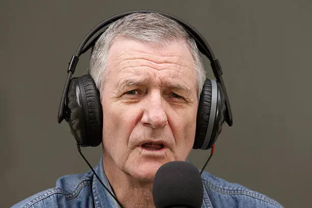 A senior radio DJ and broadcaster voices his opinion into a microphone.  He is wearing black headphones and sitting in front of a neutral grey background – ideal for replacing with a detailed backdrop.  The man is neatly groomed with short grey hair and is looking at the camera.  There is a small amount of movement blur in his lower lip as he speaks. 