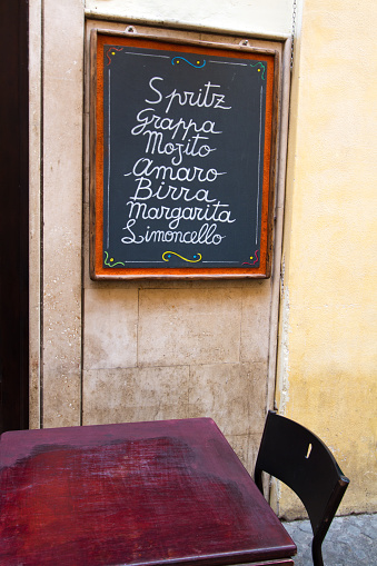 A short drink list/menu, written longhand in white chalk on a blackboard at an outdoor coffee bar in Italy. The drinks include: Spritz, Grappa, Mojito, Amaro, Birra, Margarita, and Limoncello. One (red) table is visible, as well as a yellow wall.