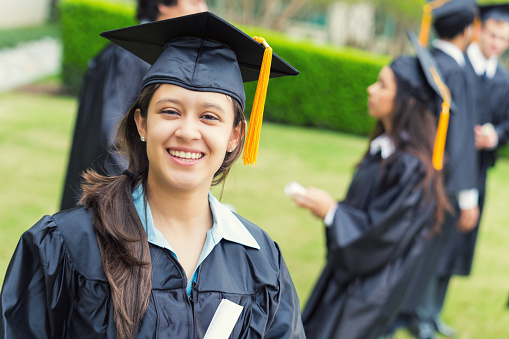 Young adult Hispanic woman is smiling. She is wearing a black graduation cap and gown and holding a diploma. Graduation cap has a gold tassle. Classmates are standing on lawn behind her after graduation ceremony. 