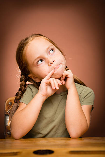 Color image of a little girl who is deep in thought while sitting at her school desk. On a brown background.