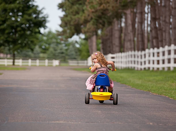 Young girl riding her tricycle and looking back over her shoulder. The girl has curly blonde/brown hair and is wearing a pink and brown dress.