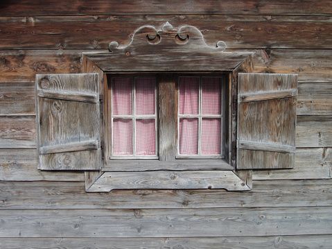 The facade of an old wooden log building with a small window divided into 4 parts and inside you can see a cup placed on a table.