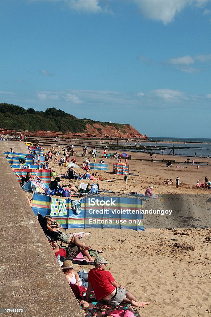 People on Exmouth Beach Exmouth, United Kingdom - August 22, 2012: Picture of Exmouth Beach in the afternoon. The picture includes the sandy beach, some people sitting down with a wall behind them and the beach in front of them. Some families and friends are using wind brakes to give them some privacy from others.There is also a cliff that dominates most of the centre left side of the picture. Adult Stock Photo
