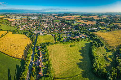 Suburban homes, gardens and factories surrounded by patchwork quilt landscape of green pasture, golden crops and wooded hills in this aerial vista. ProPhoto RGB profile for maximum color fidelity and gamut.