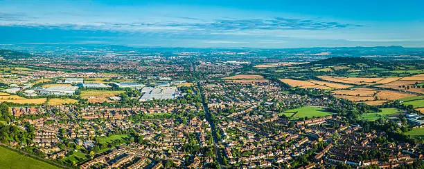 Suburban homes, gardens and factories surrounded by patchwork quilt landscape of green pasture, golden crops and wooded hills in this aerial panoramic vista. ProPhoto RGB profile for maximum color fidelity and gamut.