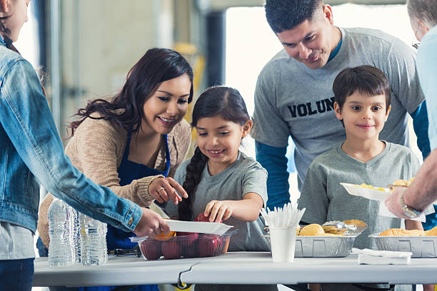 Family serving meals while they volunteer in soup kitchen together Young Hispanic family is serving healthy meals to people in charity food bank soup kitchen. Mother and father are instructing elementary age little boy and little girl as they serve food to senior man and preteen girl.  soup kitchen stock pictures, royalty-free photos & images