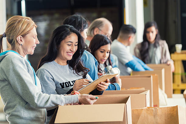 Diverse adults packing donation boxes in charity food bank Mature adult Caucasian woman is standing with mid adult Hispanic woman as they pack cardboard boxees full of donated food in charity food bank. Other volunteers are lined up behind them, also sorting donated groceries into boxes. Hispanic woman is writing on checklist on clipboard. humanitarian aid stock pictures, royalty-free photos & images