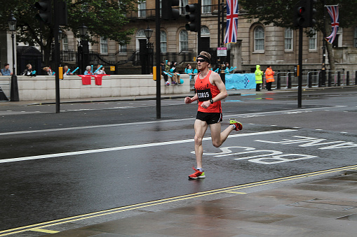 London, England - July 08, 2012: A colour photograph of a runner at the 2012 British London 10K, the lamp posts are decorated with the union jack. 300,000 runners participated and it was sponsored by Nike & Tiger Balm. The run starts at wellington arch and ends at Whitehall towards Trafalgar square. runners pass some very famous places including Big Ben Houses of Parliament, The London Eye, St Paul’s Cathedral, Trafalgar Square, Nelson’s Column and Westminster Abbey. There are some spectators to the left of the runner and two security staff in orange and green visors. The runner has a headband, sunglasses, and orange top and bleach shorts. He is running on the bike lane. 