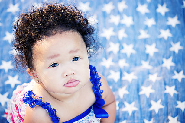 Baby Girl Looking From Up Patriotic Star Blanket Beautiful six month old multi ethnic baby girl with curly brown hair looking up at the camera. Sitting on a blue quilt with white stars and wearing a red white and blue romper. Fourth of July. patriotic quilt stock pictures, royalty-free photos & images
