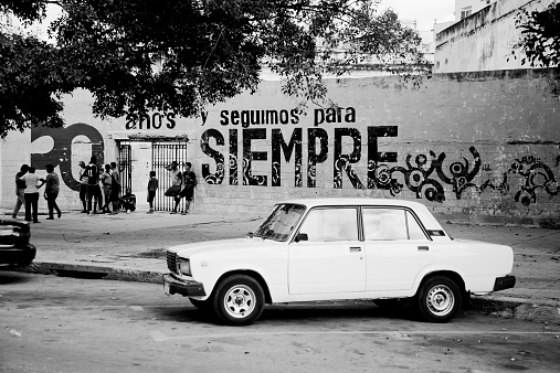 Havana, Cuba - February 23, 2010: Old Lada 2107 Russian car parked against public school with local schoolboys and propaganda slogan on the wall. With an estimated 60,000 vintage cars still in Cuba, these old classics are a tribute to the nostalgia of the old days. In monochrome.
