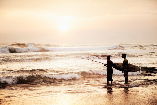 Hikkaduwa, Sri Lanka - March 9, 2013: A surfer stands next to a fisherman while he fishes in the ocean at sunset in Hikkaduwa, Sri Lanka.