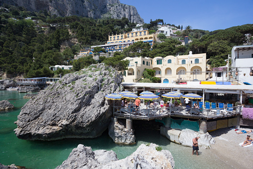 Marina Piccola, Italy - May 3, 2013: A man walks ankle deep in the water on the Marina Piccola beach while other bathers soak up the sun. Above them is a restaurant built into the rocks. 