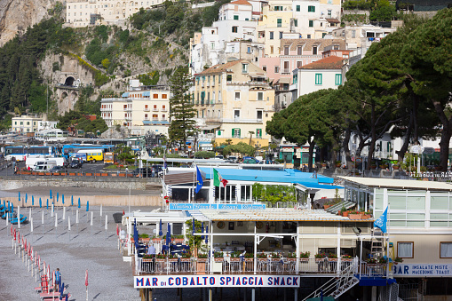 Amalfi, Italy - April 29, 2013: A view of the beach at Amalfi on the Amalfi Coast, Marina Grande, with beach snack bars in the foreground and a waiter ensuring the cutlery is in place on the table in the foreground while there are many cars and vehicles in the transport hub beyond.