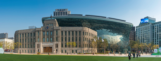 Seoul, Korea - 26th April 2013: The historic stone facade of Seoul Metropolitan Library in front of the futuristic glass wave of Seoul City Hall overlooking pedestrian strolling across Seoul Plaza, the wide green space in the heart of downtown Seoul, South Korea's vibrant capital city. Composite panoramic image created from seven contemporaneous sequential photographs.