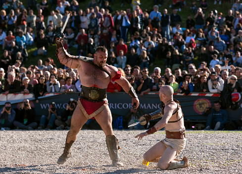 Rome, Italy - April 21, 2013: Gladiators fighting in historical costumes with ancient weapons in Circo Massimo, Rome, during Rome's Foundation Celebrations. One is standing and the other one kneels in disadvantage, defending himself from the blow. Gladiators' fighting was one of the most required entertaining activity. This event has been organized by Gruppo Storico Romano and involved 11 different countries, 2000 people and 53 different volunteer associations. All the costumes are historically correct thanks to the supervision of a team of archeologists and researchers. The leading volunteer group of Gruppo Storico Romano was born in 1994. By now, the group has highly increased and has a scientific department of experimental archeology which expresses itself by recreating ancient situations such as parties or gladiators' fights and dances.