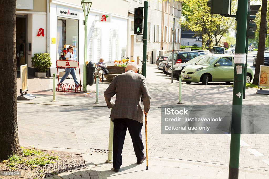 Old in town Essen, Germany - May, 6th 2013: Senior man with walking stick is crossing traffic light on Rüttenscheiderstraße in Essen Bredeney. In background is sitting another senior on bench in front of pharmacy. A young man is walking on sidewalk. Active Seniors Stock Photo