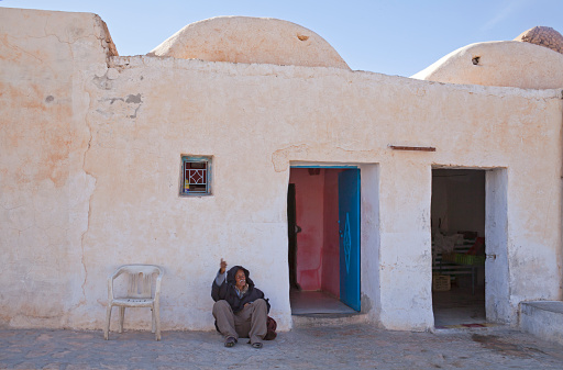 Douz, Tunisia, March 30, 2013: An old man wearing a brown tunic is sitting on the floor beside a chair in front of a row typical houses, looking and pointing up