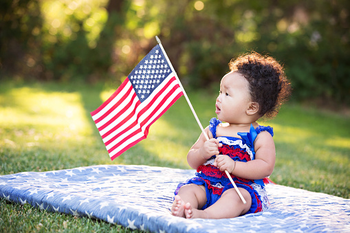 Beautiful six month old multi ethnic baby girl sitting on a blanket at the park and looking at the American flag she is holding. 4th of July.