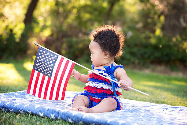 Baby Girl Sitting and Holding American Flag Beautiful six month old multi ethnic baby girl sitting on a blanket at the park and holding an American flag. 4th of July. patriotic quilt stock pictures, royalty-free photos & images