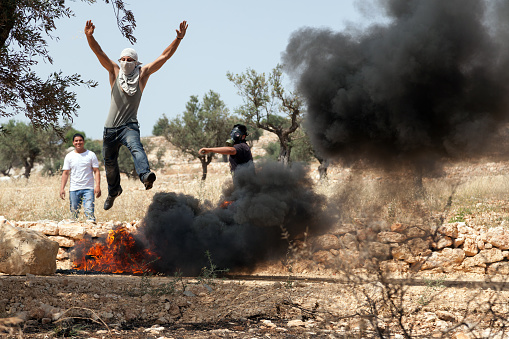 Bil'in, Israel - May 17th, 2013: A young Palestinian protester jumping over flaming tires at a protest against the Israeli occupation, with his friends watching him.