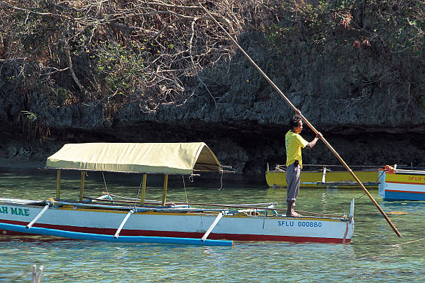 Filipino Boat Man, Hundred Islands Pangasinan, Philippines- February 01, 2013: A colour photograph of a Filipino man wearing a yellow T-shirt and Khaki pants holding on to a long peace of bamboo/ stick that he is using to row his traditional boat. In the background is the base of on of The hundred Islands. pangasinan stock pictures, royalty-free photos & images