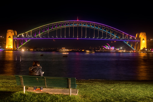Sydney, Australia - May 25, 2013: A woman watches from Blues Point Reserve as the Sydney Harbour Bridge and Opera House are colourfully lit up. These projections are part of the annual Vivid Sydney festival, with the Harbour Bridge being lit up for the first time on this occasion.