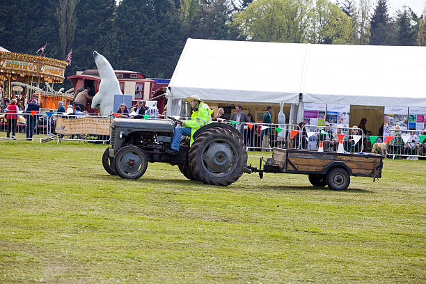 Old vintage diesel Tractor and Trailer Henley on Thames, England - May 19, 2013: Old vintage diesel Tractor and Trailer: Old vintage diesel Tractor and Trailer at a Vintage Transport Rally: Behind is a Marquee, Merry-go-round and vintage fair a large Polar Bear and spectators. The driver has a yellow coat. ursus tractor stock pictures, royalty-free photos & images