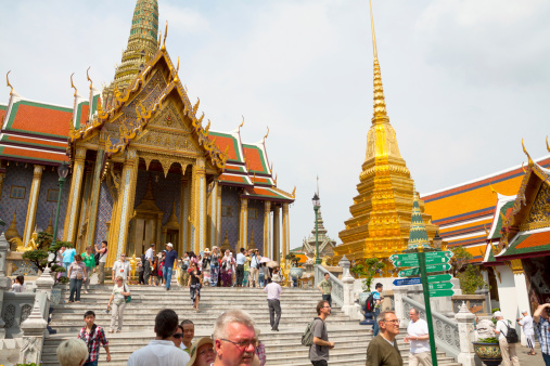 Bangkok, Thailand - February, 19th 2013: Tourists are doing sight seeing at Royal Pantheon in famous WatPhra Kaeo. They are walking upstairs and downstairs steps inf front of temple. On top are many people. At rigth side is golden pagoda with warrior statues around.