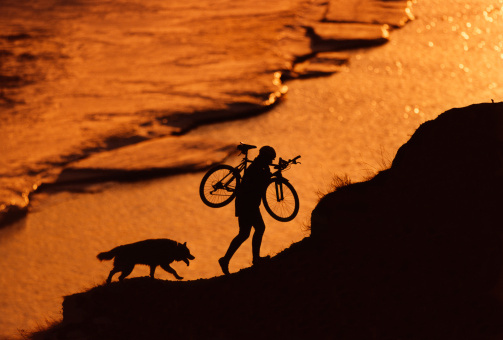 A man carries his mountain bike up a steep hill while out riding with his dog next to a flowing river partially covered in ice.