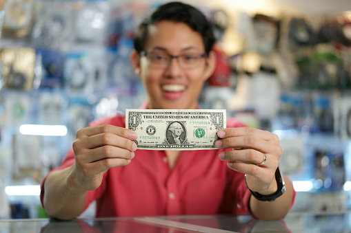 Young asian shop owner in computer store, showing his first dollar to the camera and smiling. Focus on banknote