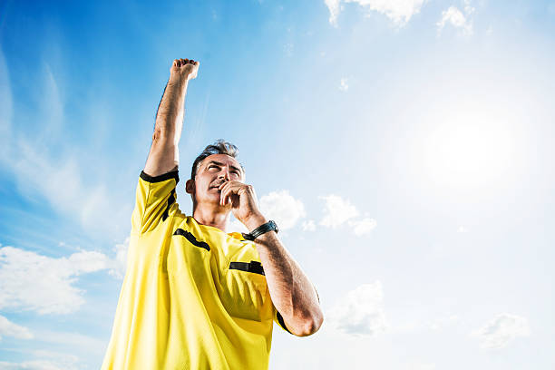 Soccer referee blowing his whistle against the sky. Low angle view of referee raising his arm and blowing his whistle against the sky. referee stock pictures, royalty-free photos & images