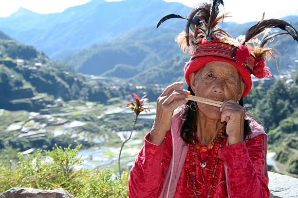 Banaue, Philippines - January 14, 2006: Old Ifugao woman in traditional clothing and headress playing a musical instrument high above the worl heritage rice terraces built into the steep mountain sides and maintained by subsequent generations of ifugao. Ifugao can be roughly translated as peple of the hill, and is also used to refer to the region in Northern Luzon.