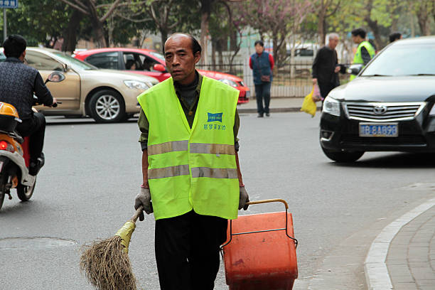 Jiangxi, China - March 10, 2013: Street cleaner working on a busy street in Jiangxi province whilst wearing a high- Vis top, carrying a broom and dragging a bin.