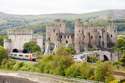 Conwy, Wales - June 2, 2013: Conwy Castle and a Virgin Train travelling through the welsh countryside. The railway embankment meets the sea and is covered with trees and shrubs but you can still see the Virgin Trains service as it passes the medieval castle at Conwy on the North Wales coast. The flag of wales a welsh dragon flutters in the wind from the top of the castle walls. To the right a road bridge with cars and on the walls of the Castle people can be seen.