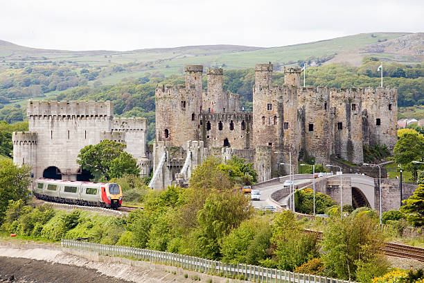 Virgin Voyager train passes Conwy Castle Conwy, Wales - June 2, 2013: Conwy Castle and a Virgin Train travelling through the welsh countryside. The railway embankment meets the sea and is covered with trees and shrubs but you can still see the Virgin Trains service as it passes the medieval castle at Conwy on the North Wales coast. The flag of wales a welsh dragon flutters in the wind from the top of the castle walls. To the right a road bridge with cars and on the walls of the Castle people can be seen. conwy castle stock pictures, royalty-free photos & images