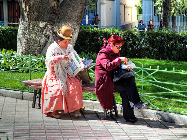 La Paz, Bolivia - May 31, 2013: Two women on public bench in the Bolivian city of La Paz. One in traditional dress is reading the day's newspaper whilst the other is eating a snack. There are three people pictured in the background.