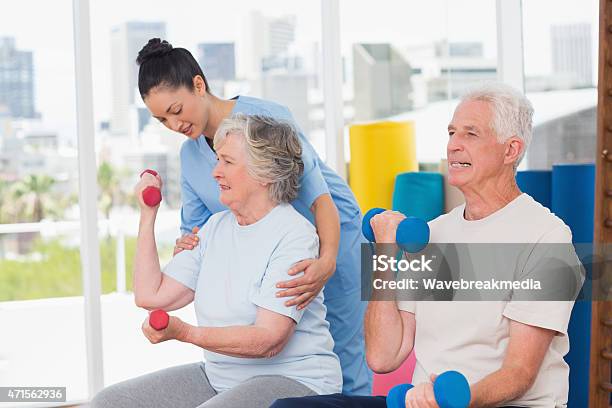 Instructor Assisting Senior Woman In Lifting Dumbbells By Man Stock Photo - Download Image Now