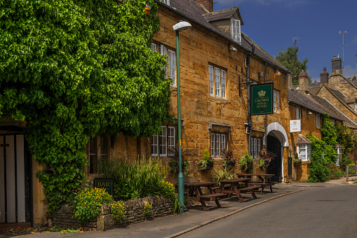 Blockley, UK - June 7, 2013: This picture shows the high street of the Cotswold village of Blockley, in the English Cotswolds, Gloucestershire, The Midlands, England, UK. The Crown Inn and Hotel is in the centre of the village, which is a local pub, offering accommodation, food and drink. It is a summer afternoon and there is intermittent sunshine lighting the honey coloured stone of the buildings. There are no people in the picture.