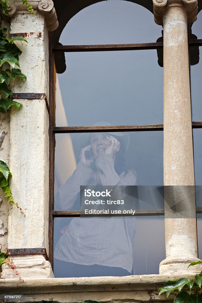Woman on mobile through old window Bled, Slovenia - July 2, 2012: Woman tourist in sunhat talking on mobile phone inside an old building through a smoked glass window Adult Stock Photo