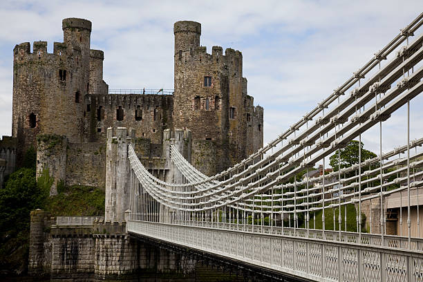 Foot Bridge to Conwy Castle Conwy, Wales - June 2, 2013: Foot Bridge leading to Conwy Castle; the stone walls of the castle towers and battlements of this medieval castle stand on the top of a hill overlooking the town. The bridge is a Victorian iron suspension bridge painted grey. A woman can be seen on the wall of the castle and a child in one of the windows.  conwy castle stock pictures, royalty-free photos & images