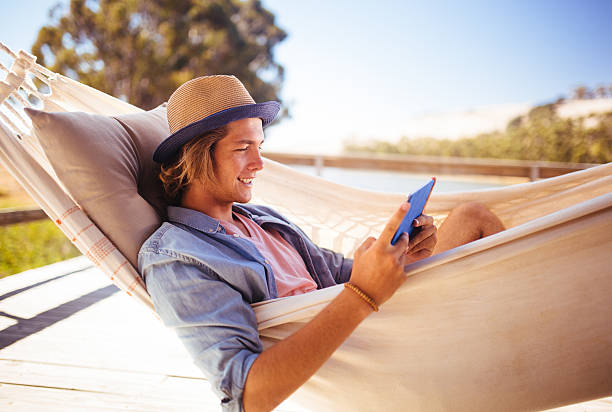 Teen reading on his digital tablet relaxing in a hammock Teen guy relaxing in a hammock while reading on his digital tablet hammock men lying down digital tablet stock pictures, royalty-free photos & images