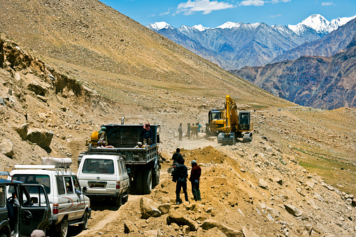 Khardung La, Jammu and Kashmir, India - July 20th, 2011:Indian road workers with machinery,  people, one on motorbike, and some cars waiting for road to be cleared, two Indians sitting on a truck. Indian mountainous roads are constantly being repaired or reconstructed because of weather and geological conditions.