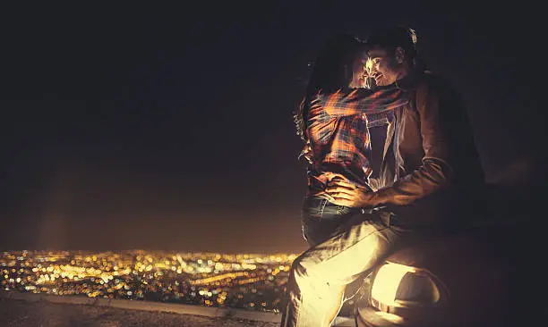 Young couple kissing on the bonnet of a car with the city lights in the background at night