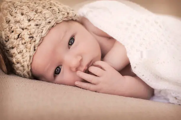 A newborn baby with his fingers in his mouth, wearing a light brown knit hat.  The baby is lying on his right side on a beige blanket and has open, dark eyes.  A white knit blanket covers the baby's bare shoulders.