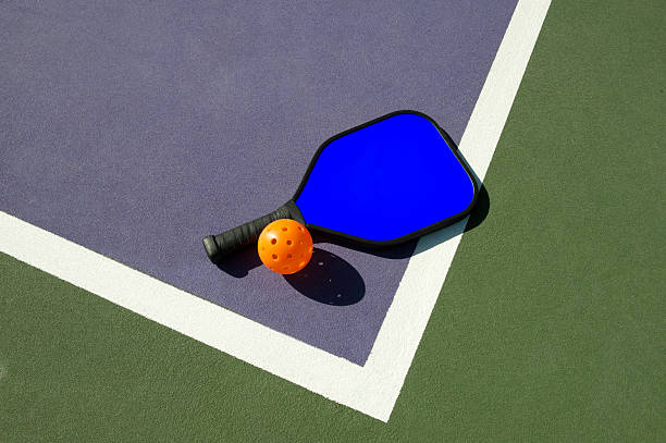 Pickleball and Blue Paddle on Edge of Court Simple geometric image of a pickleball and a pickleball paddle laying on the edge of a colorful pickleball court. table tennis bat stock pictures, royalty-free photos & images