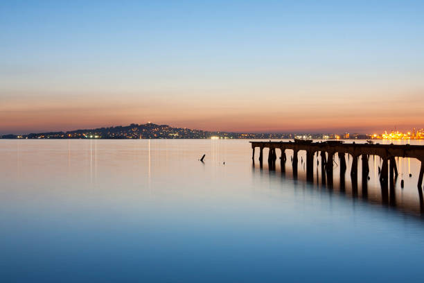 A DSLR photo of a Montevideo cityscape. View of Montevideo's harbor bay at dusk with blurred water. An abandoned pier stands on the right in diagonal pointing to the center. The \