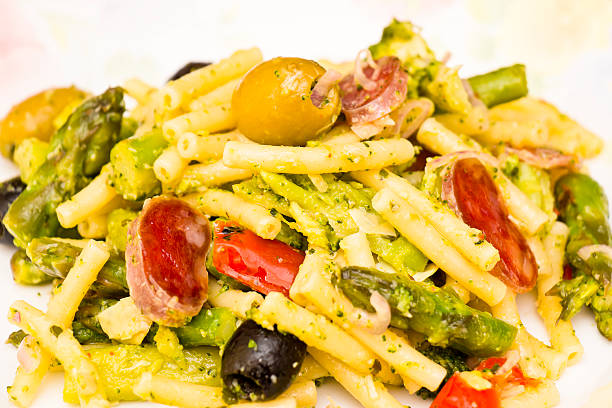 Close up of a portion Italian pasta salad with made of ziti noodles, green asparagus, broccoli, green and black olives, onions, grilled peppers, pepperoni sausage. Served with olive oil, lemon juice and grated parmesan cheese.